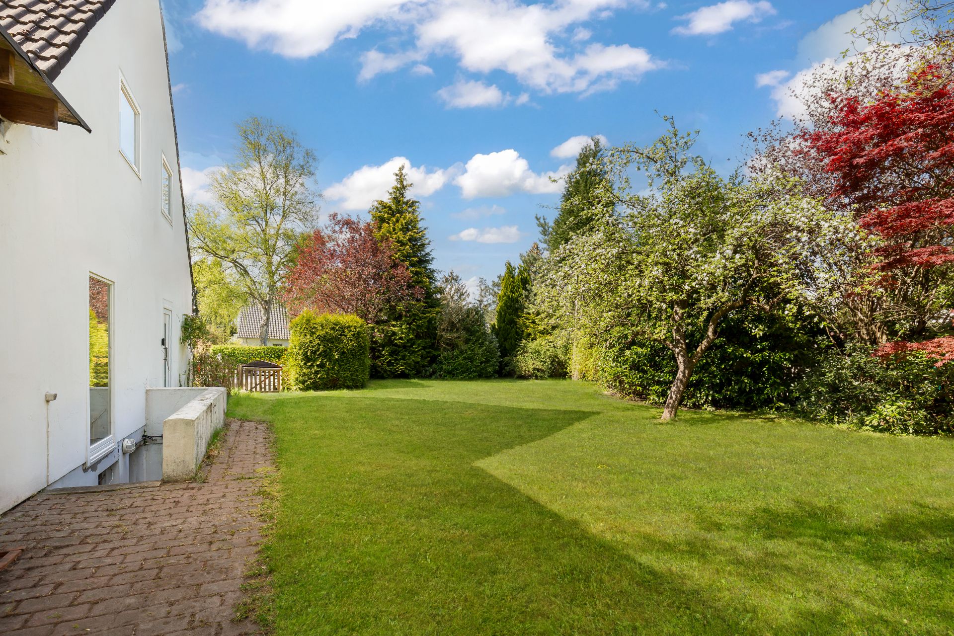 Grüner Garten mit Bäumen und Rasen neben einem weißen Wohnhaus an einem sonnigen Tag.
