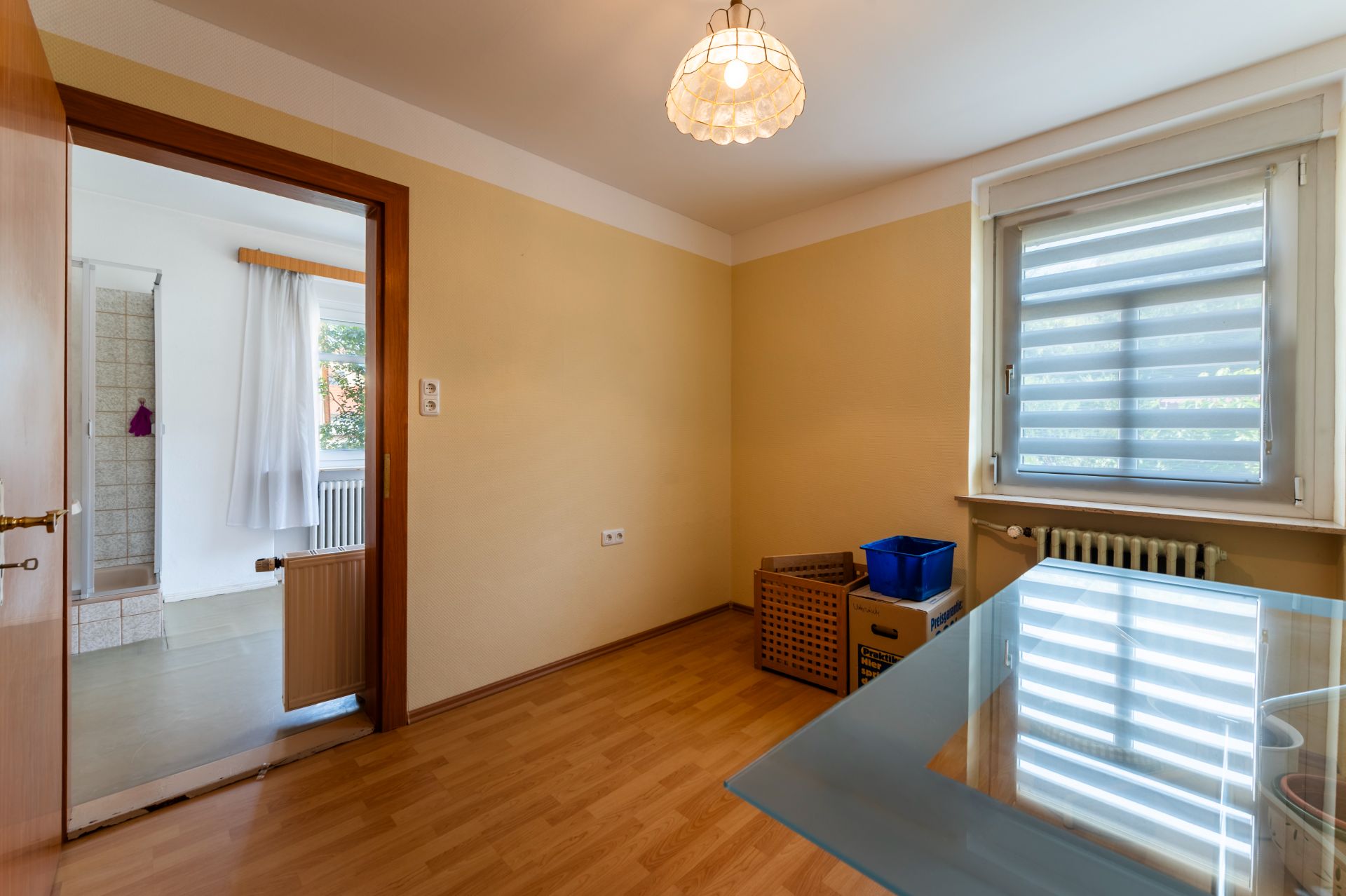 Helles Zimmer mit Holzfußboden, Glastisch, Fenster mit Jalousie und offenen Tür zur Dusche.
