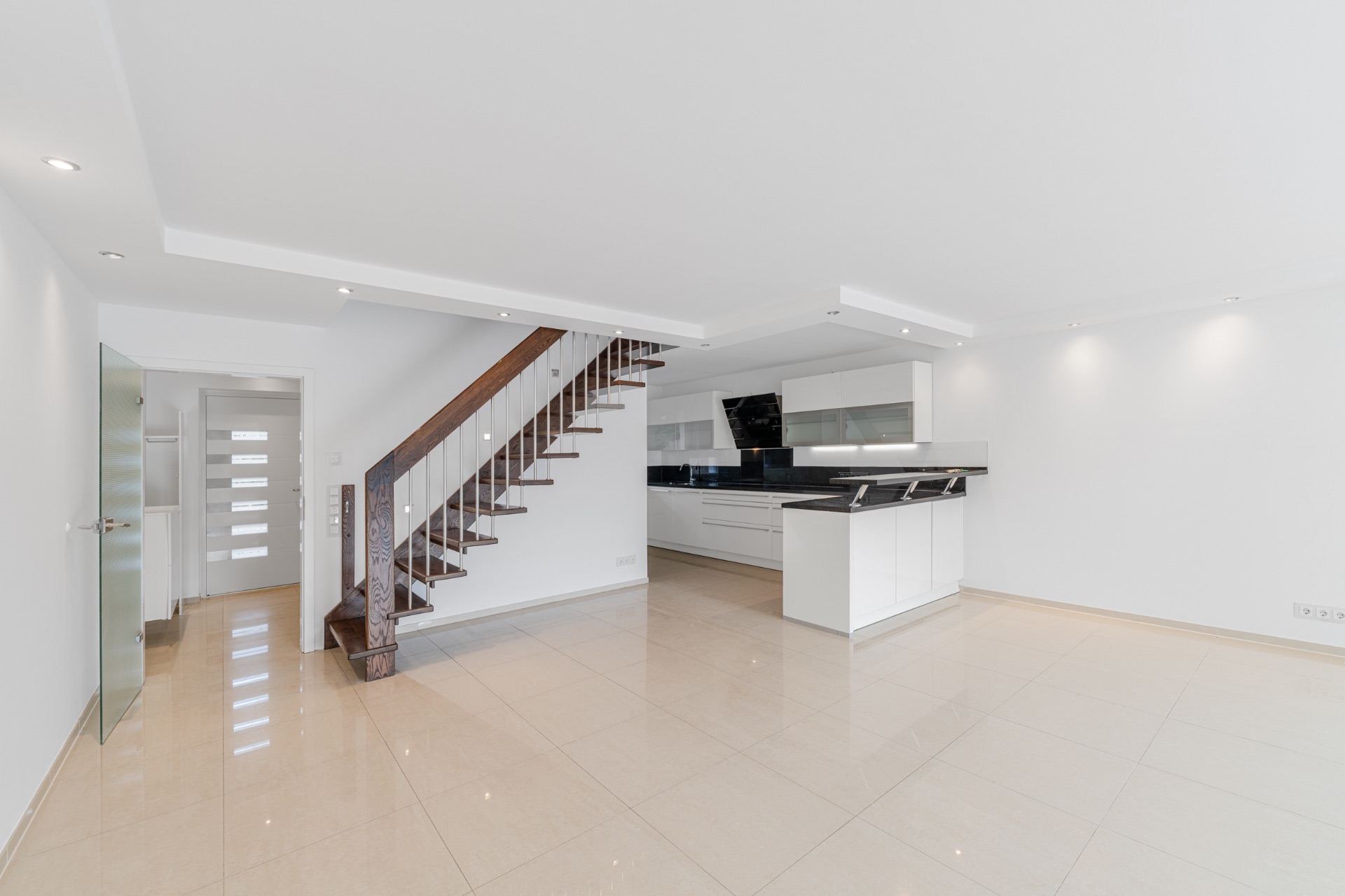 Heller, moderner Wohnraum mit offener Küche und Holztreppe, Fliesenboden.