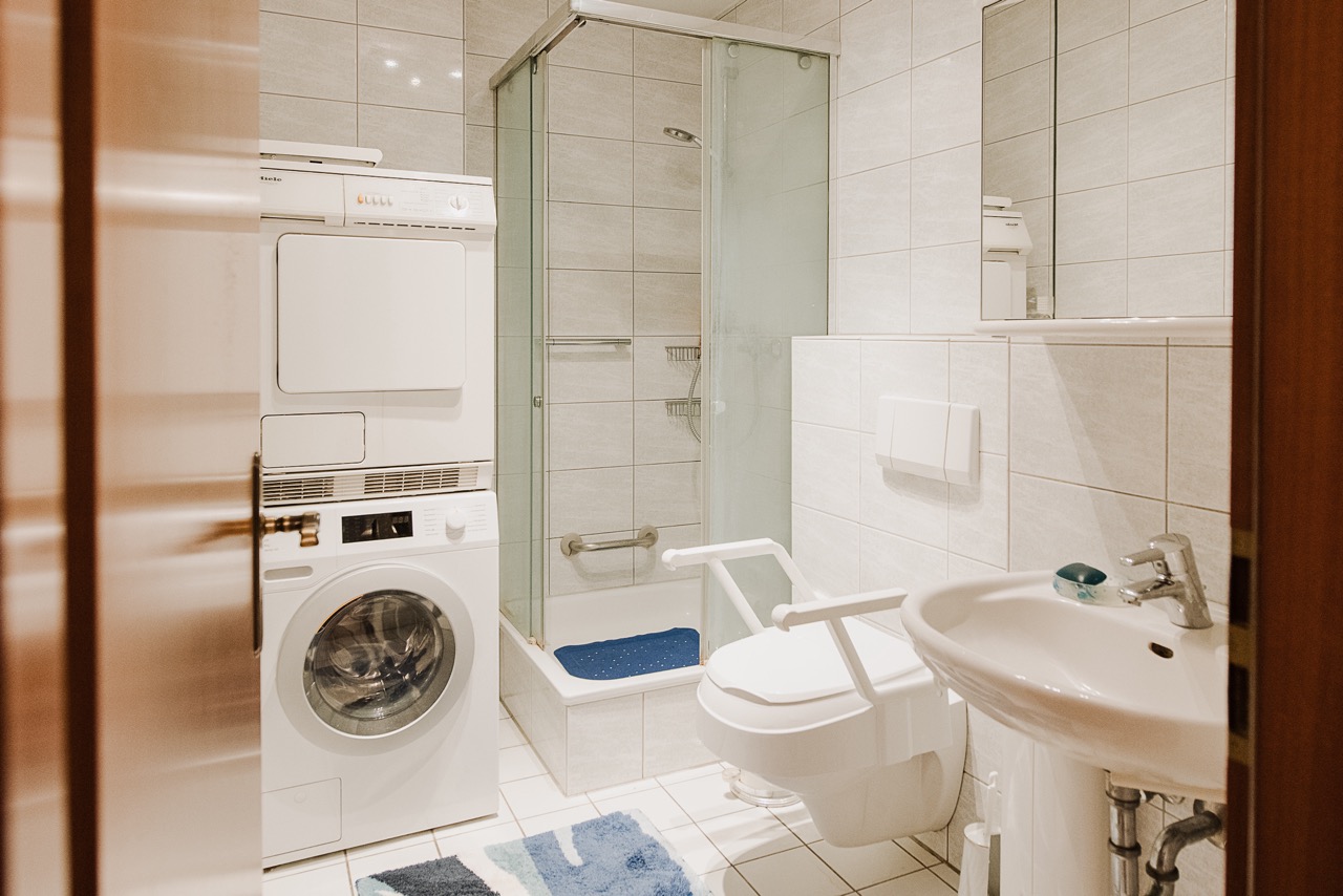 Helles Bad mit Dusche, Waschmaschine, Trockner und Toilette mit Haltegriffen.