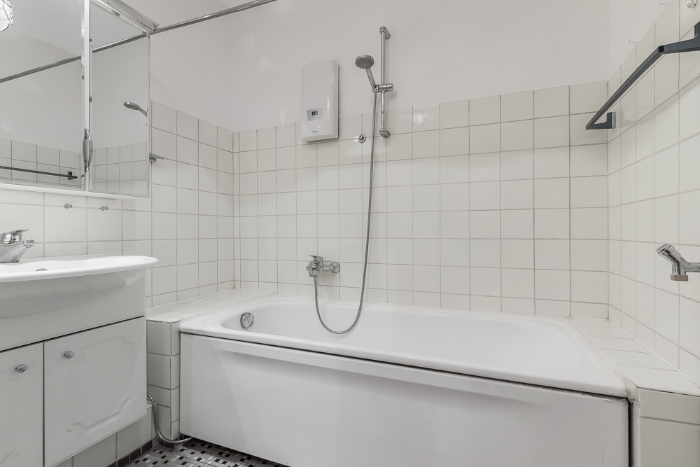 Weiß gefliestes Badezimmer mit Badewanne, Spiegel, Waschbecken und Duschkopf.