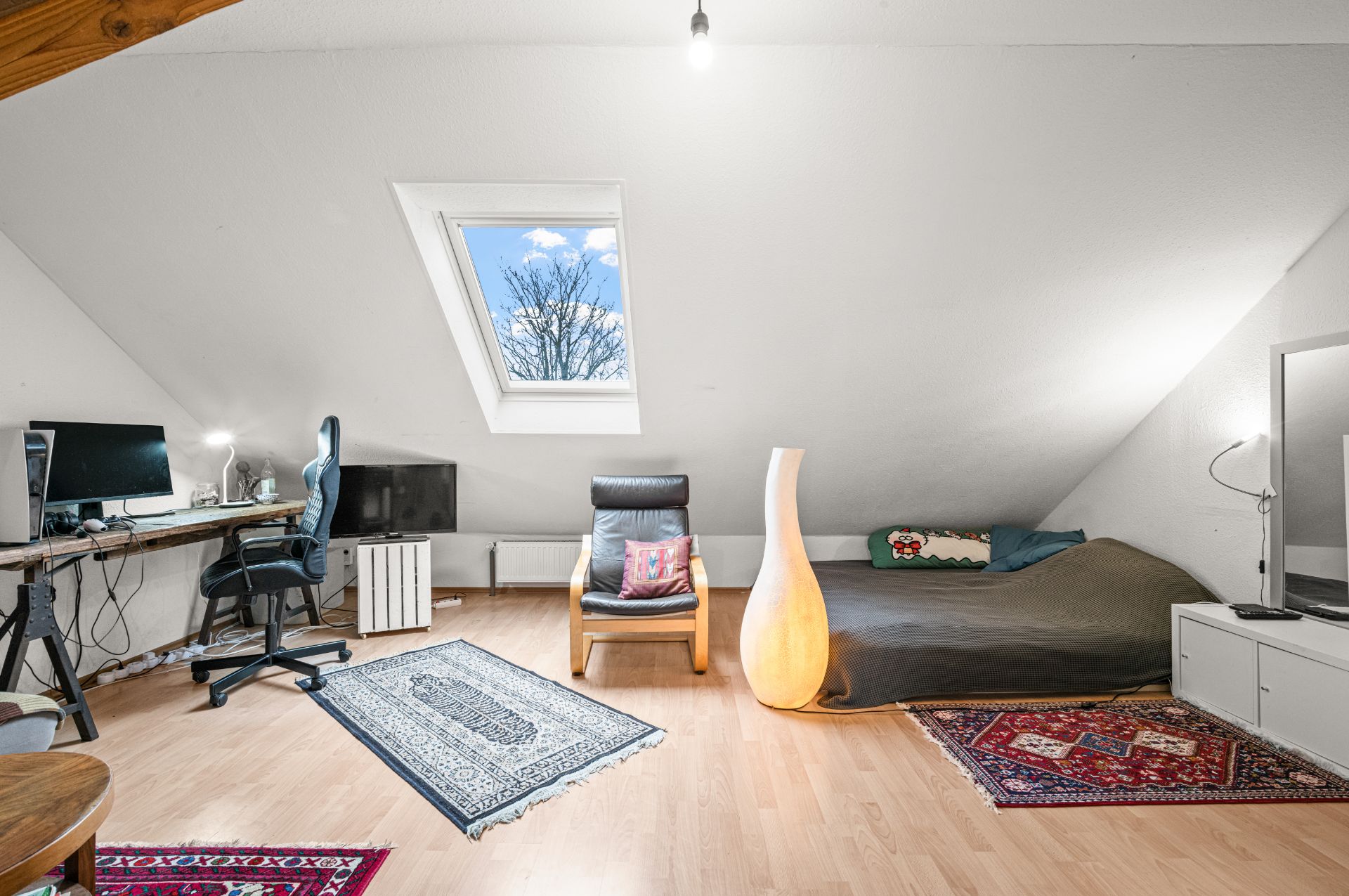 Dachzimmer mit Schreibtisch, Stuhl, Sessel, Bett, Teppichen und Dachfenster.
