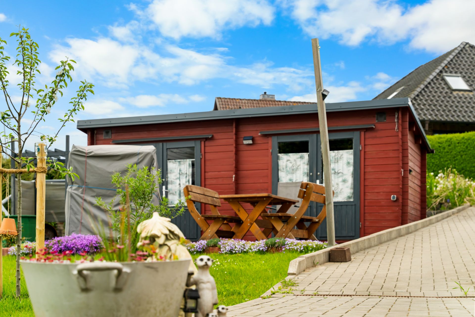 Rotes Gartenhaus mit Holztisch und Stühlen auf einer Terrasse, umgeben von grünem Rasen und Blumen.
