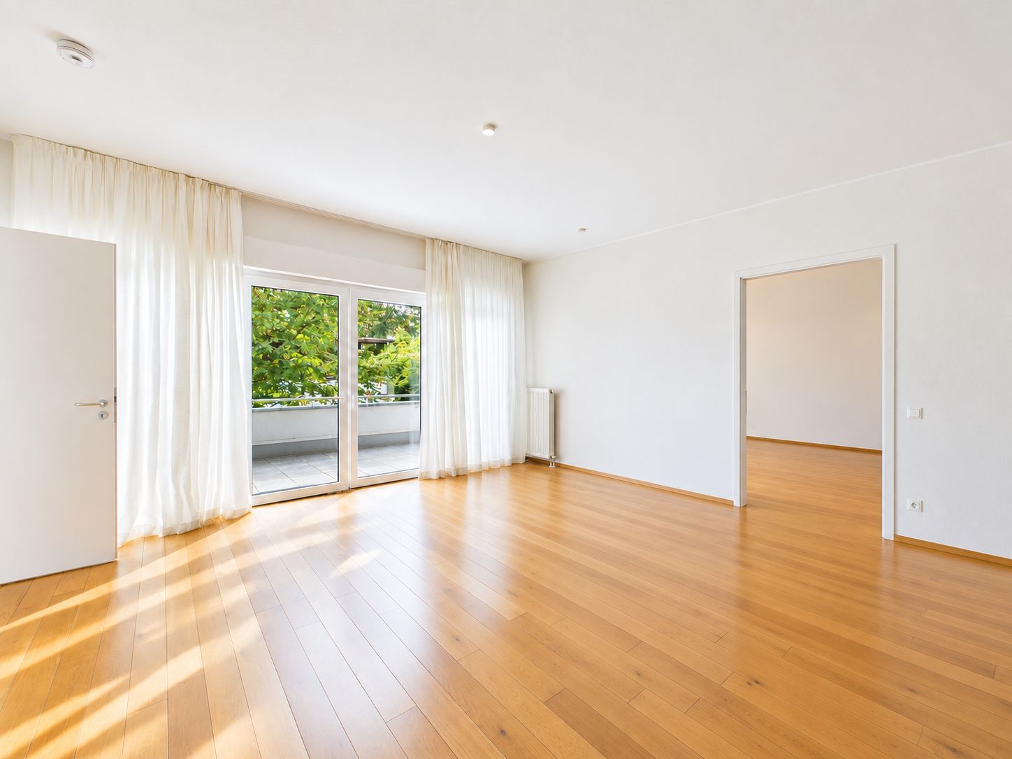 Helles, leeres Zimmer mit Holzfußboden, großen Fenstern und Zugang zu einem Balkon.