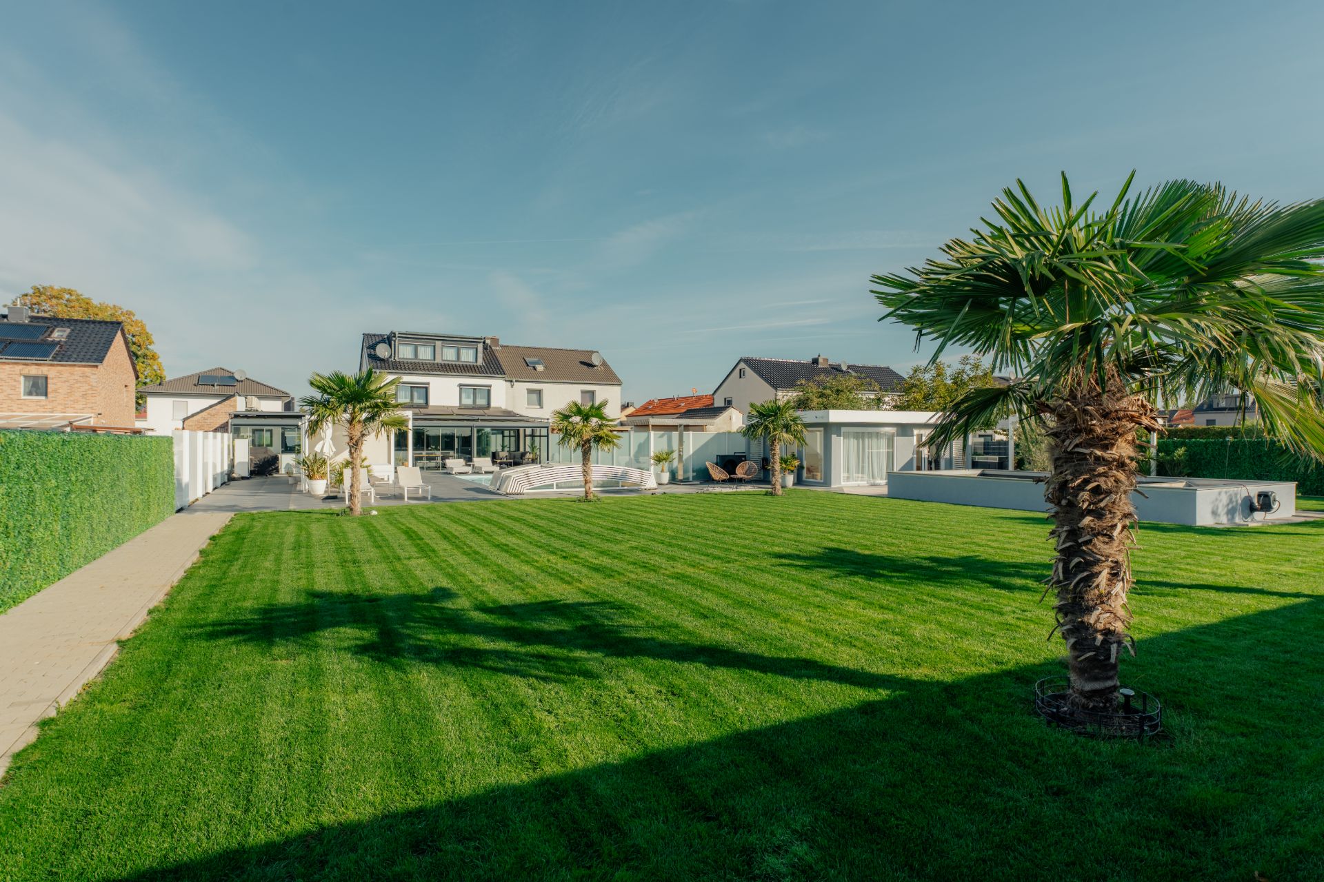 Großer Garten mit Rasenfläche, mehreren Palmen und modernen Wohnhäusern im Hintergrund.