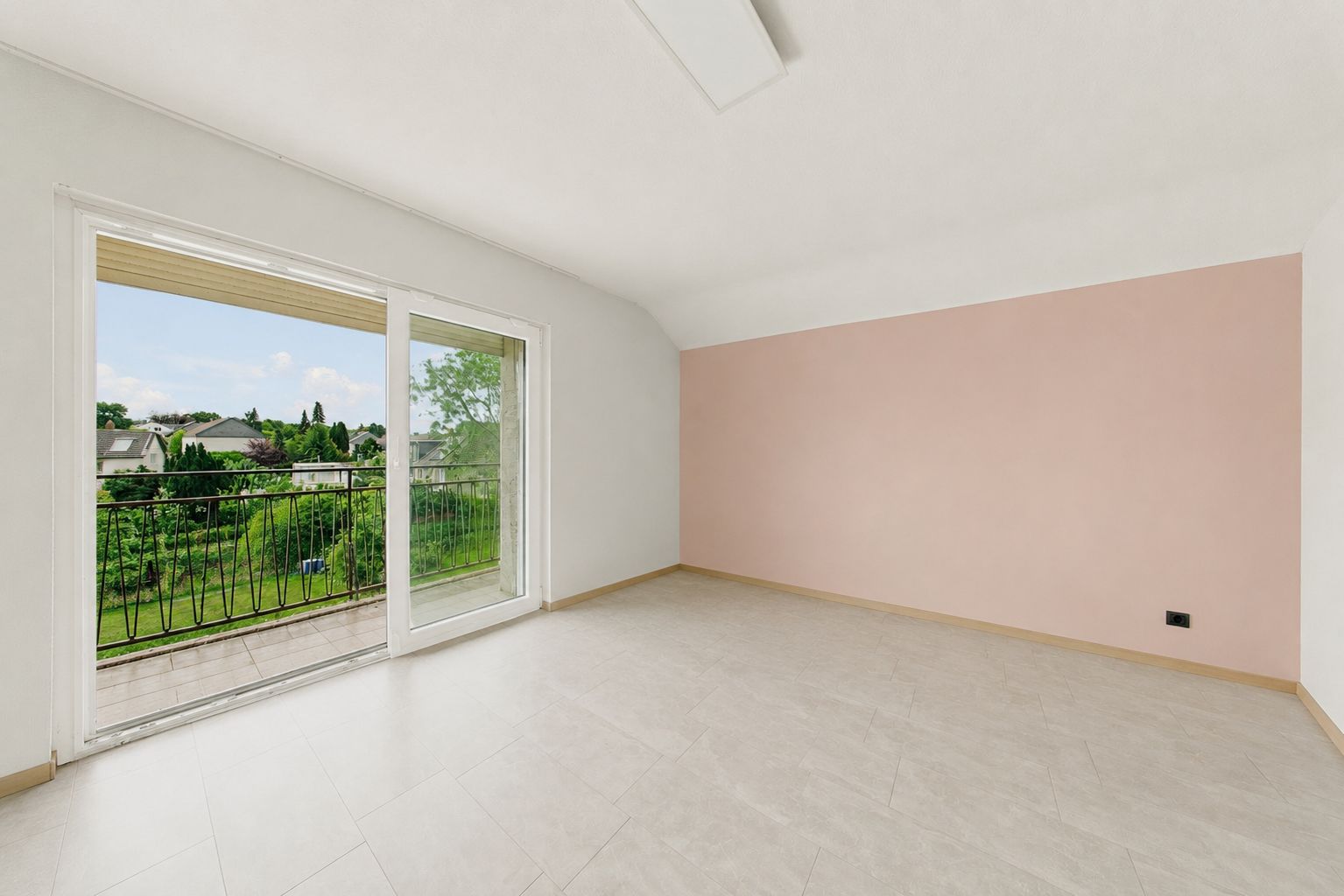 Leerer, heller Raum mit rosa Wand und großer Glastür zum Balkon mit Blick ins Grüne.