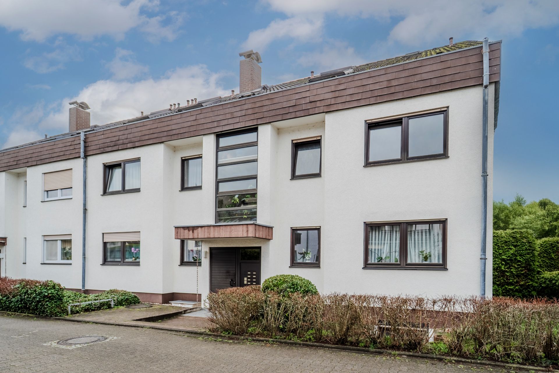Mehrfamilienhaus mit weißer Fassade, braunem Dach und mehreren Fenstern, umgeben von Sträuchern.