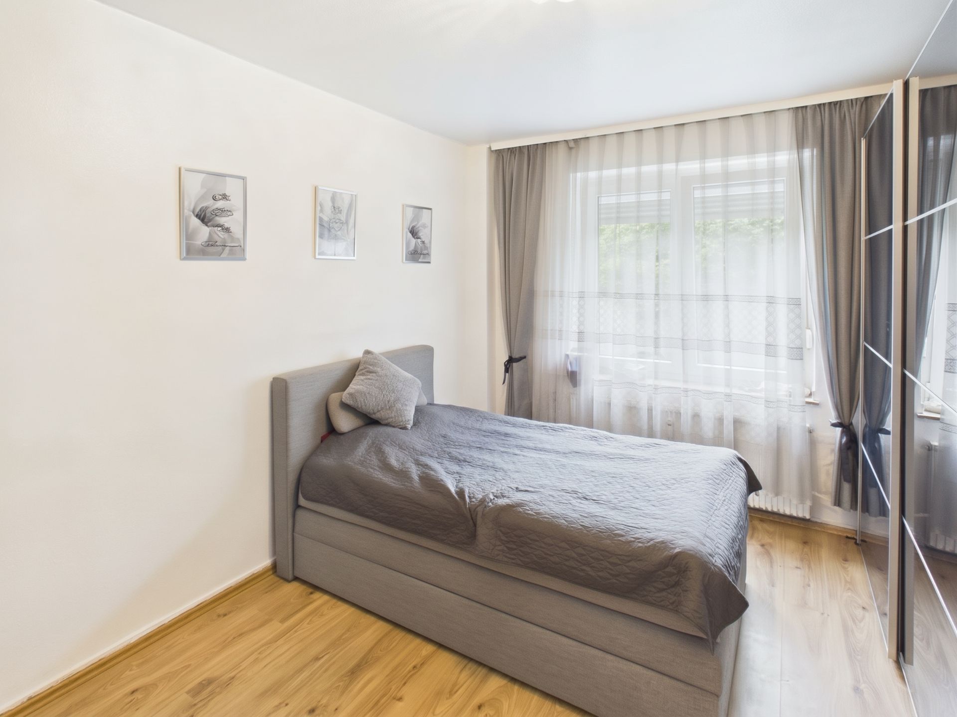Helles Schlafzimmer mit Einzelbett, grauer Bettwäsche und großem Fenster mit Vorhängen.