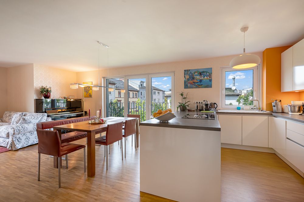 Helle Wohnküche mit Esstisch, Sofa, Kücheninsel und großen Fenstern mit Ausblick.