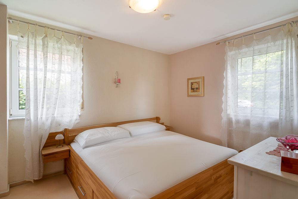 Helles Schlafzimmer mit Doppelbett, zwei Fenstern mit Vorhängen und Nachttischlampe.