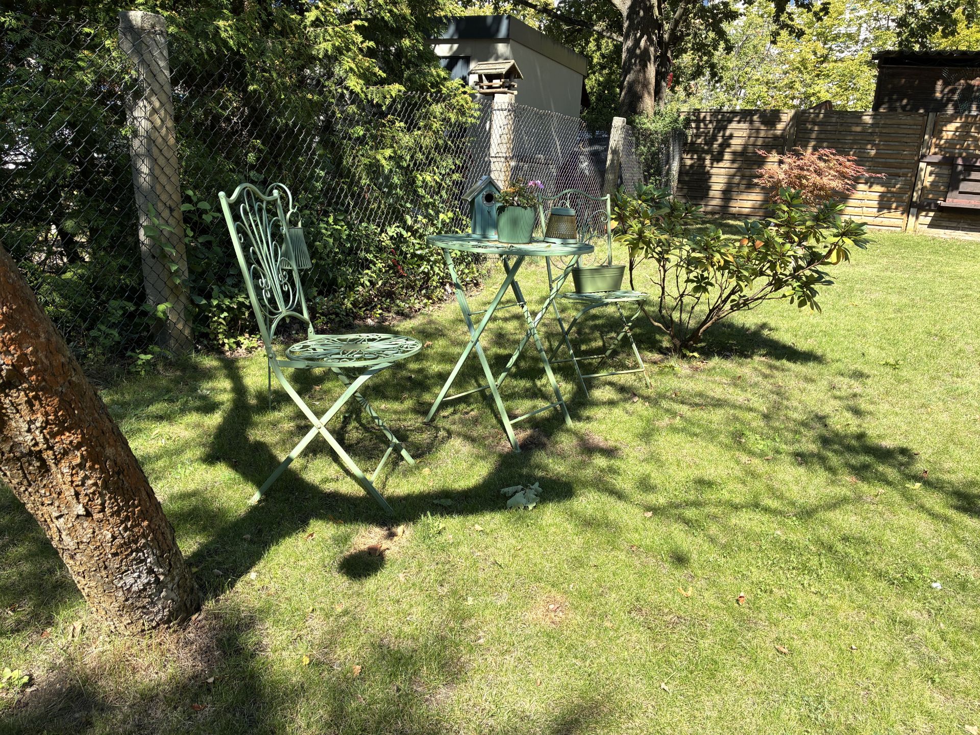 Zwei grüne Gartenstühle und ein Tisch mit Blumentöpfen stehen auf einer sonnigen Wiese.