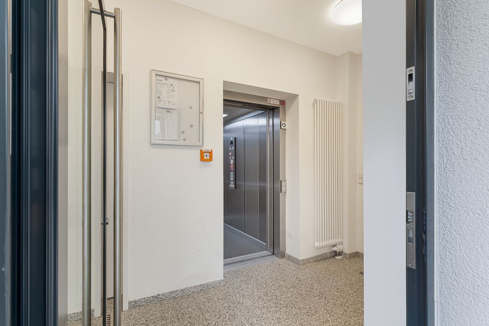 Offener Aufzug in einem hellen Flur mit Glastür und Wandheizkörper.