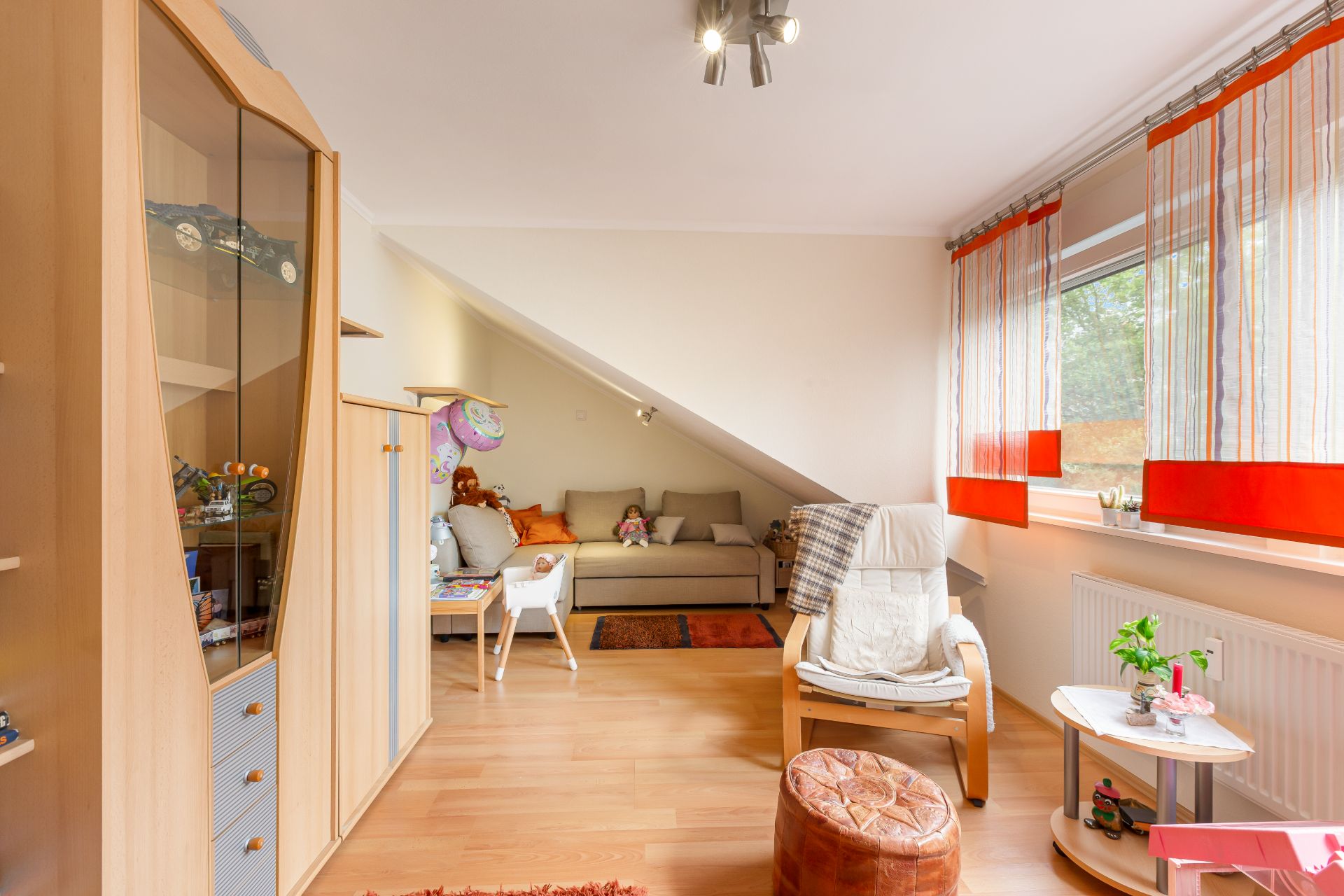 Helles Kinderzimmer mit Sofa, Sessel, Spielzeug und großem Fenster mit roten Gardinen.