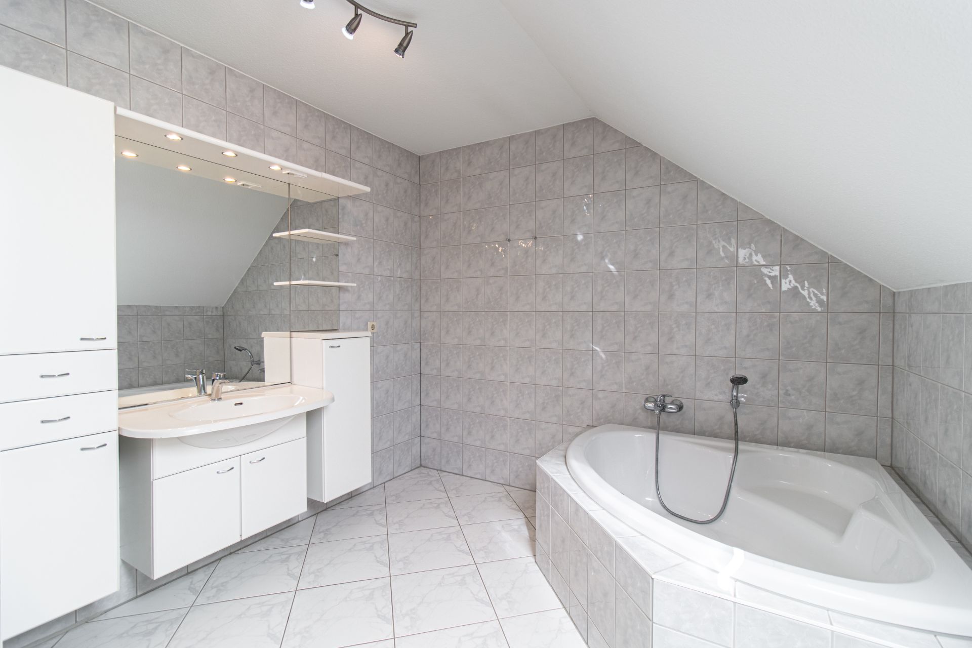 Modernes Badezimmer mit Eckbadewanne, großem Spiegel und weißen Schränken unter Dachschräge.