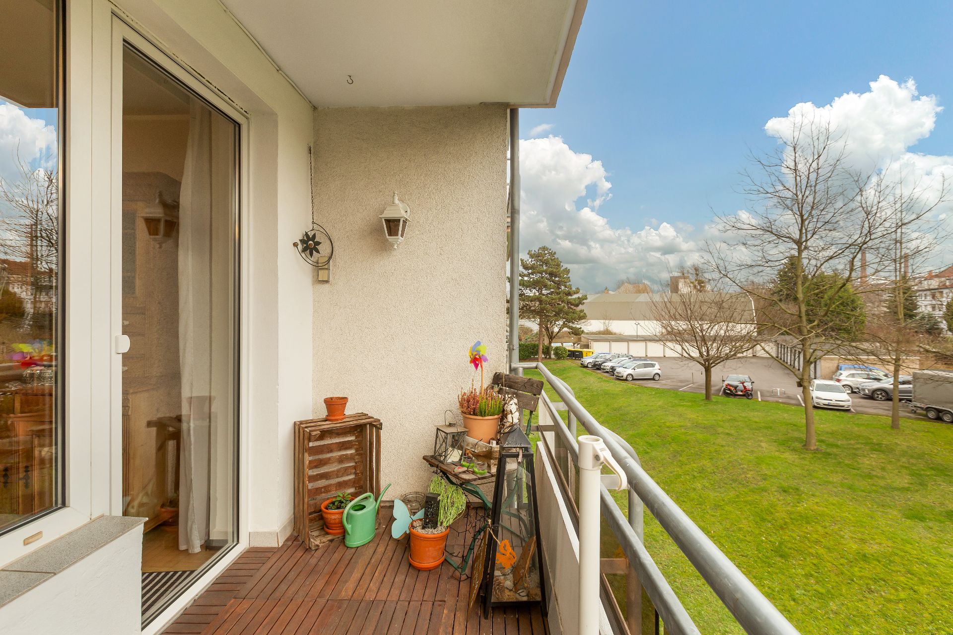 Balkon mit Holzboden, Pflanzen, Gießkannen und Blick auf eine grüne Wiese und einen Parkplatz.