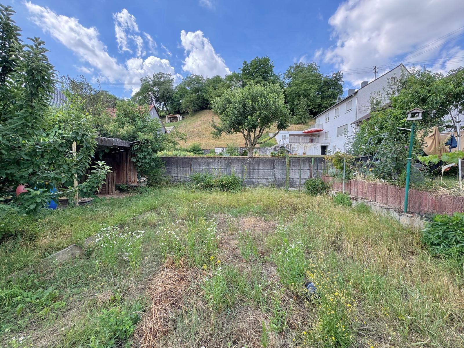Garten mit Gras, Bäumen und Holzschuppen, im Hintergrund Wohnhäuser und Hügel.