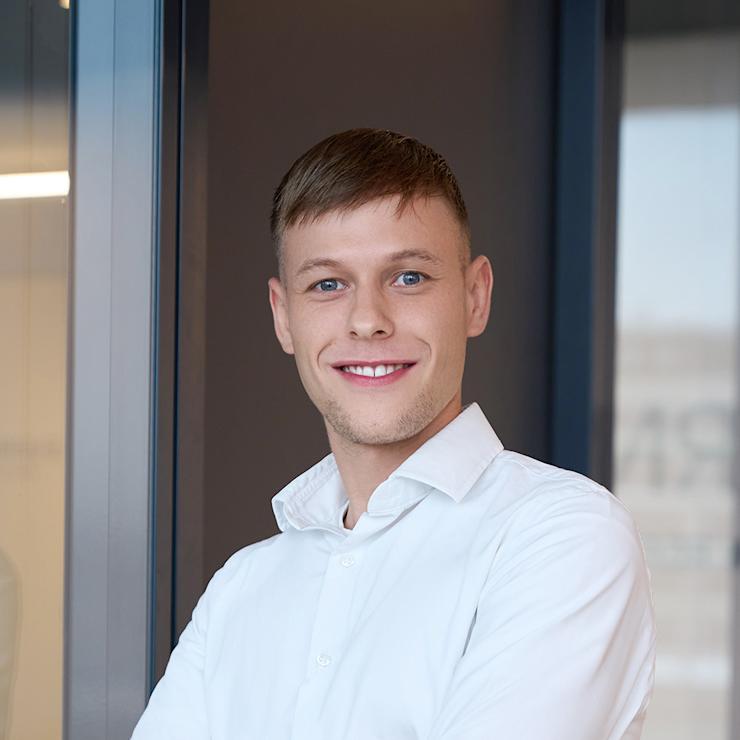 Junger Mann mit weißem Hemd steht lächelnd mit verschränkten Armen vor Glaswand.