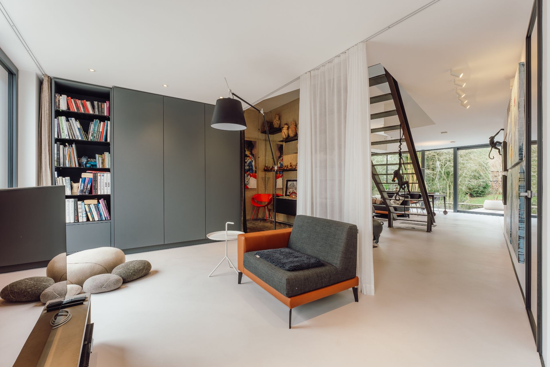 Modernes Wohnzimmer mit Bücherregal, Sessel, Stehlampe und Treppe zu einem hellen Bereich mit Fenstern.