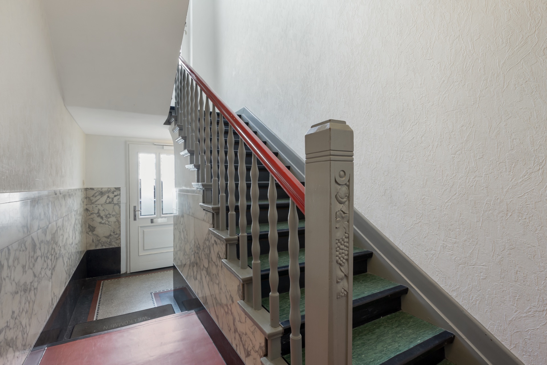 Treppenhaus mit Marmorelementen und rotem Handlauf, Tageslicht durch Glastür.