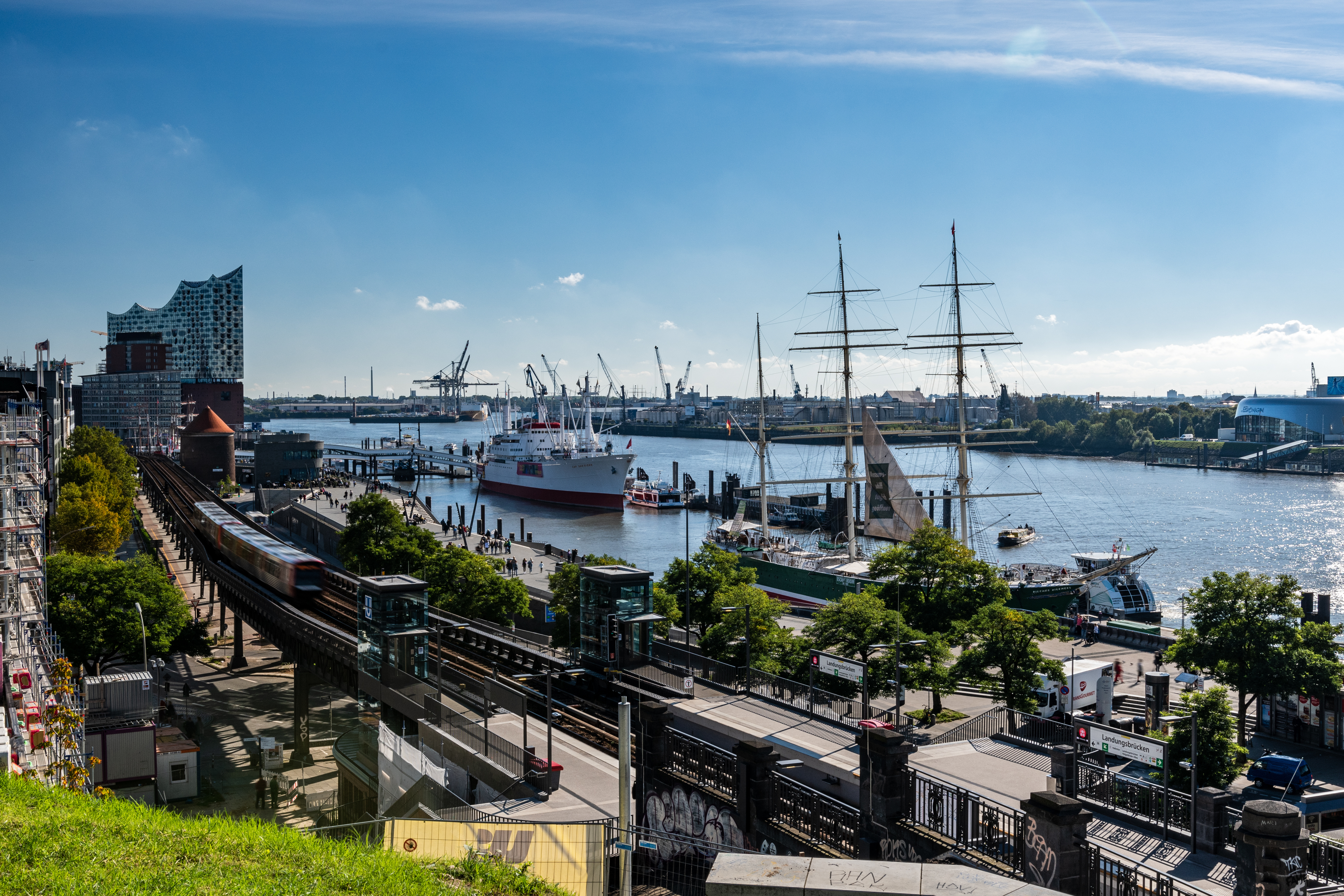 Aussicht auf einen Fluss mit Schiffen, einem Zug und modernen Gebäuden im Hintergrund.