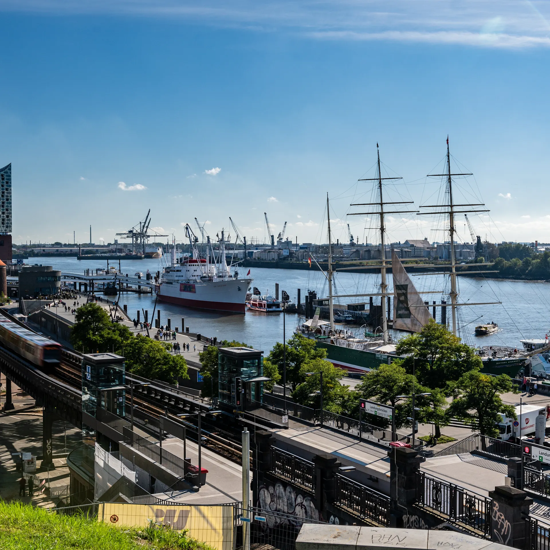 Aussicht auf einen Fluss mit Schiffen, einem Zug und modernen Gebäuden im Hintergrund.
