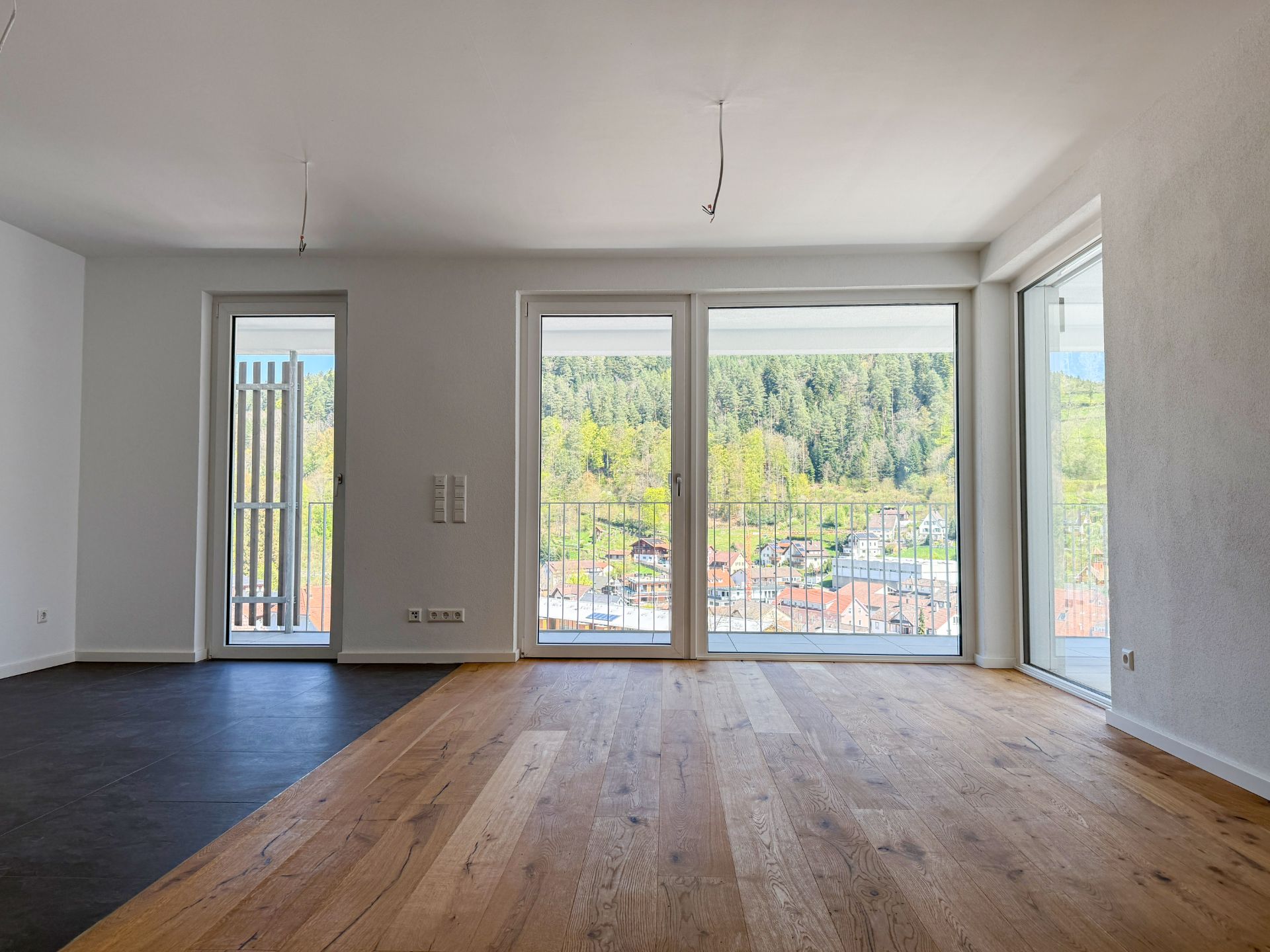 Helles Zimmer mit großen Fenstern, Holzboden und Ausblick auf einen Wald und eine Stadt.