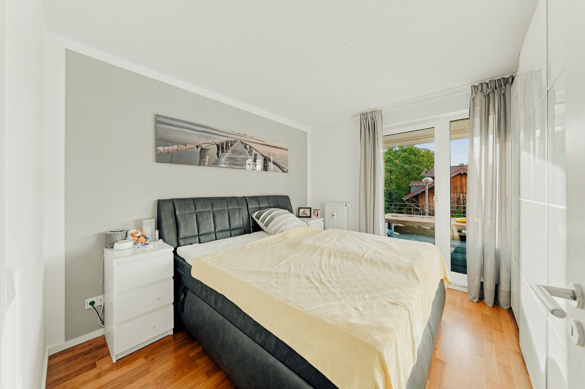 Helles Schlafzimmer mit großem Bett, Nachttisch, Fenster und Vorhängen, Holzboden.