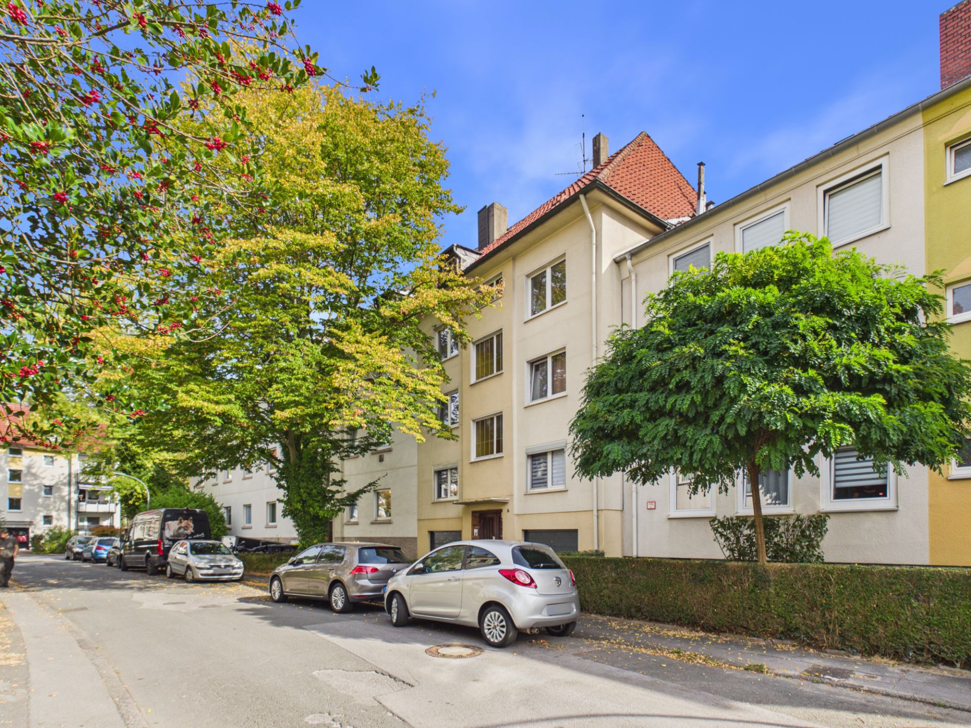 Mehrfamilienhaus mit Bäumen und geparkten Autos an einer ruhigen Wohnstraße.