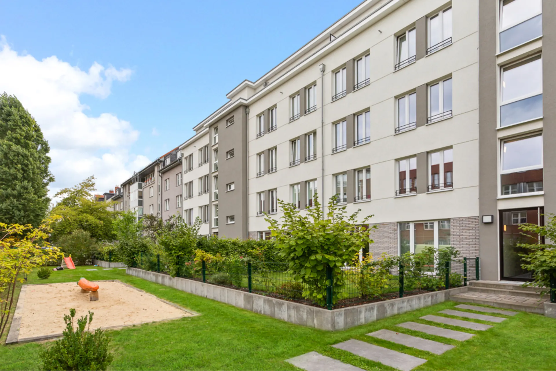 Mehrstöckiges Wohngebäude mit Garten, Sandkasten und Rutsche im Vordergrund.