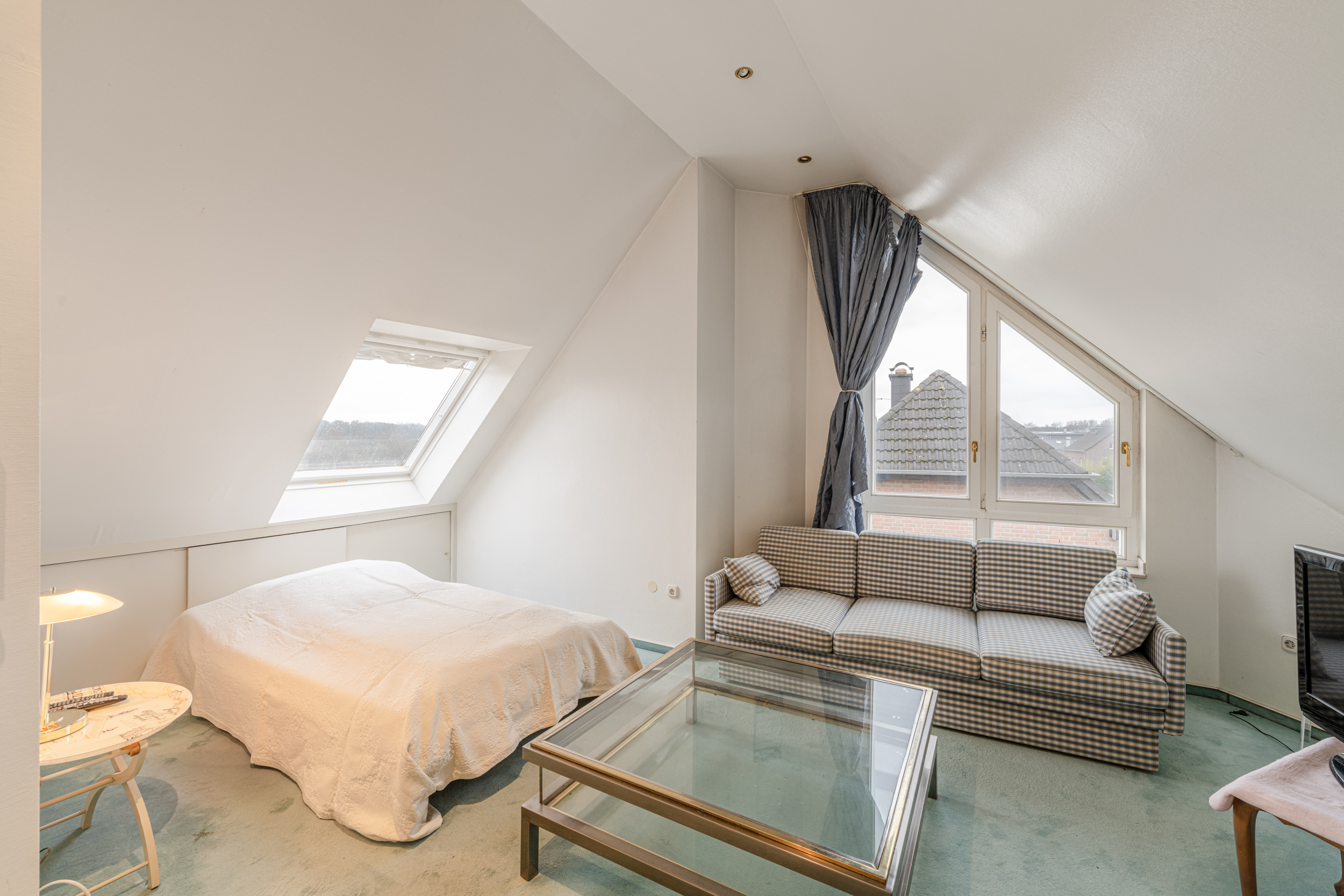 Helles Zimmer mit Bett, kariertem Sofa, Glastisch und großen Fenstern im Dachgeschoss.