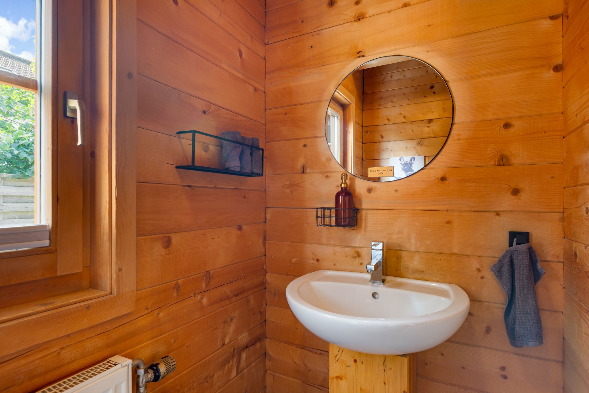 Kleines Badezimmer mit Holzverkleidung, Waschbecken, rundem Spiegel und Fenster.