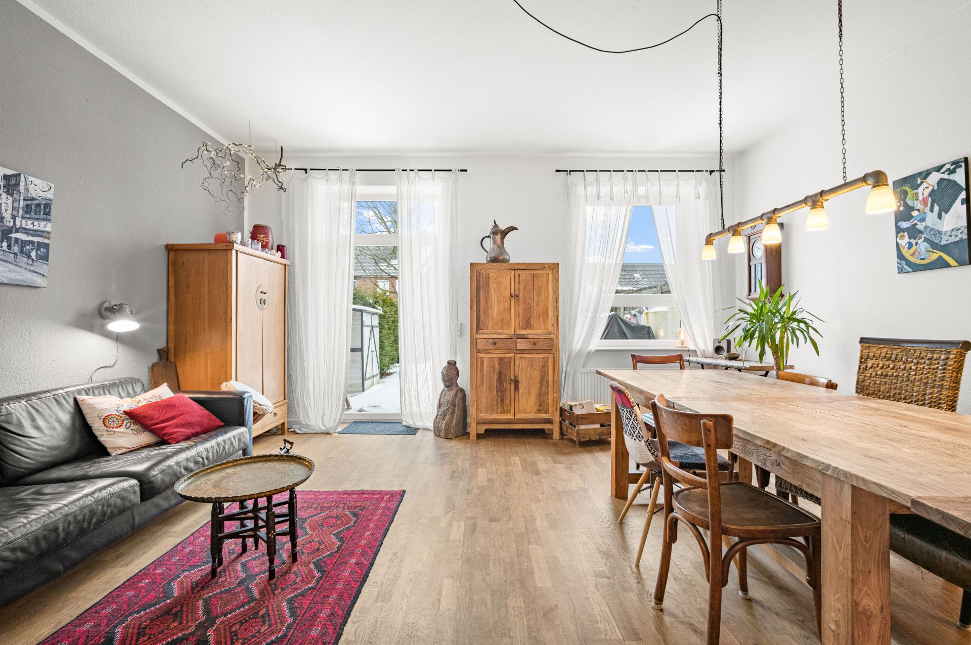 Helles Wohnzimmer mit Holzboden, Sofa, Esstisch, Schrank und großen Fenstern mit weißen Vorhängen.
