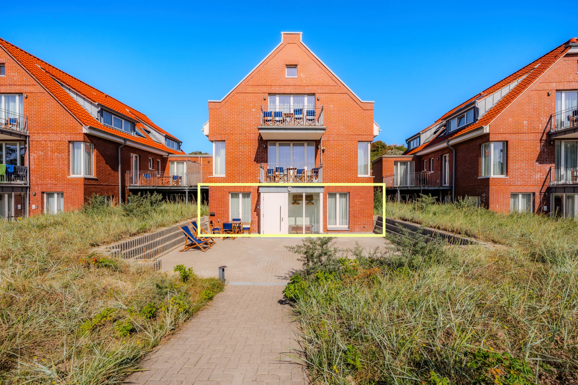 Rotes Backsteinhaus mit Terrasse, Balkon, Liegestühlen und Gartenweg im Vordergrund.