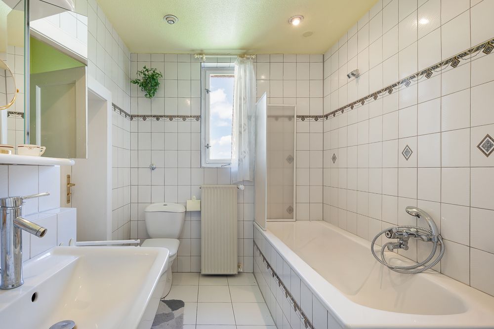 Helles Badezimmer mit Badewanne, Waschbecken, WC und Fenster, alles mit weißen Fliesen.