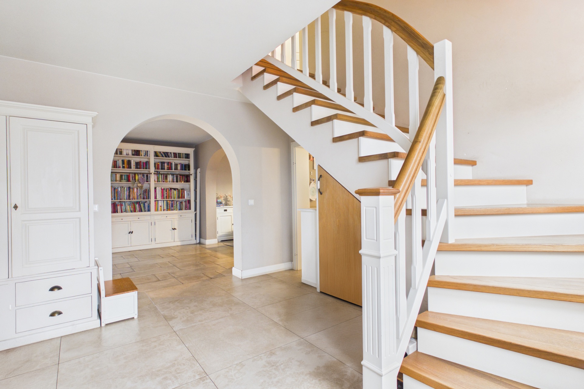 Heller Flur mit Holztreppe, weißem Schrank und Regal mit Büchern durch einen Torbogen sichtbar.