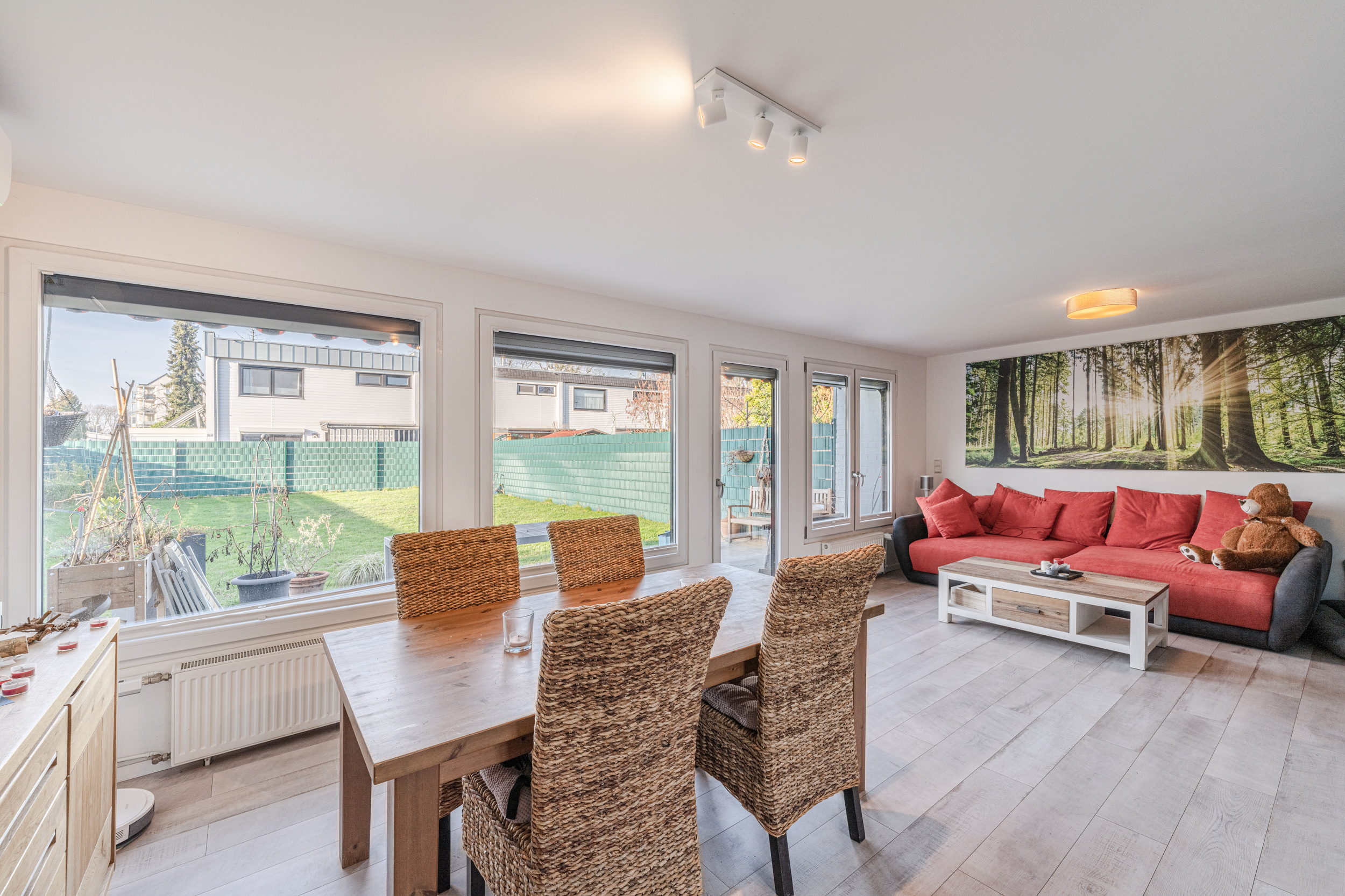 Helles Wohnzimmer mit großem Fenster, Esstisch, Korbstühlen und rotem Sofa mit Wandbild.