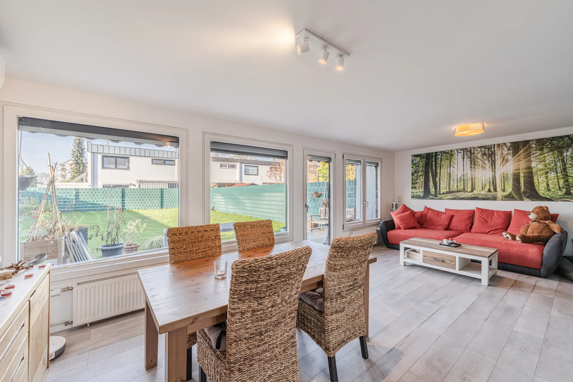 Helles Wohnzimmer mit großem Fenster, Esstisch, Korbstühlen und rotem Sofa mit Wandbild.