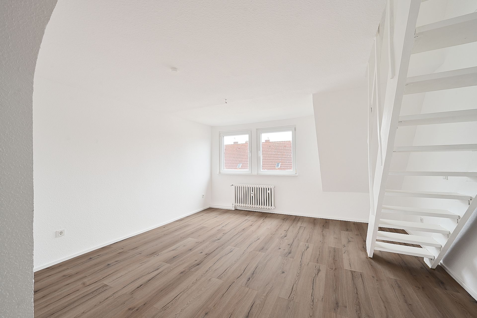 Helles, leeres Zimmer mit Holzfußboden, zwei Fenstern und einer weißen Treppe.