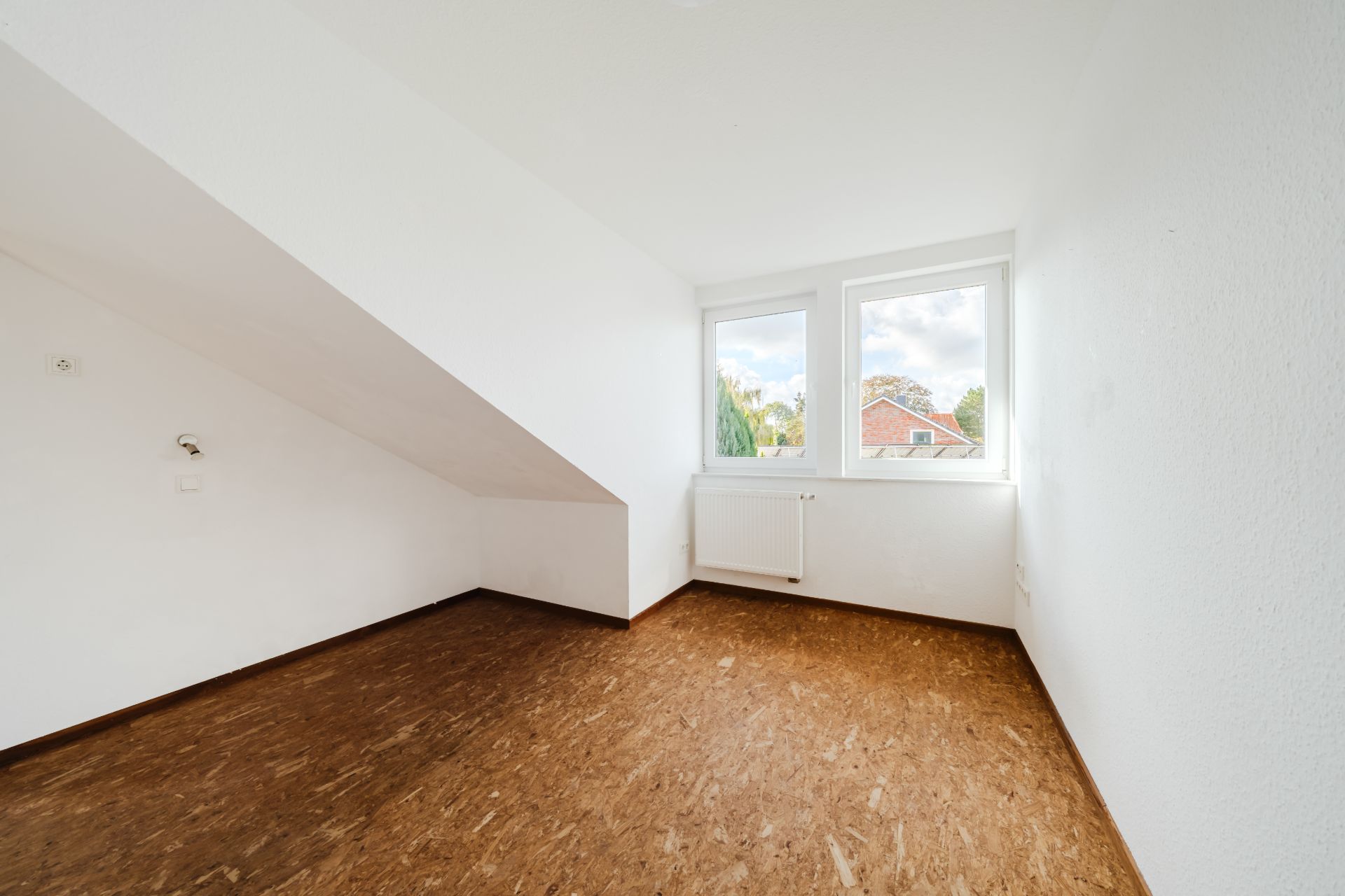 Leerstehendes Zimmer mit schräger Decke, zwei Fenstern und Heizkörper, brauner Boden.