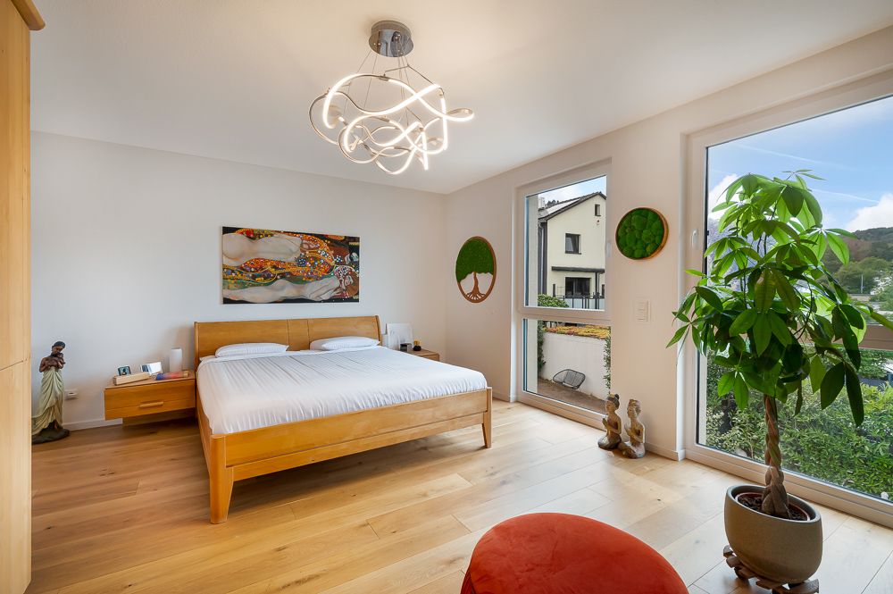 Helles Schlafzimmer mit Holzfußboden, Doppelbett, großer Pflanze und Fenstern mit Blick nach draußen.