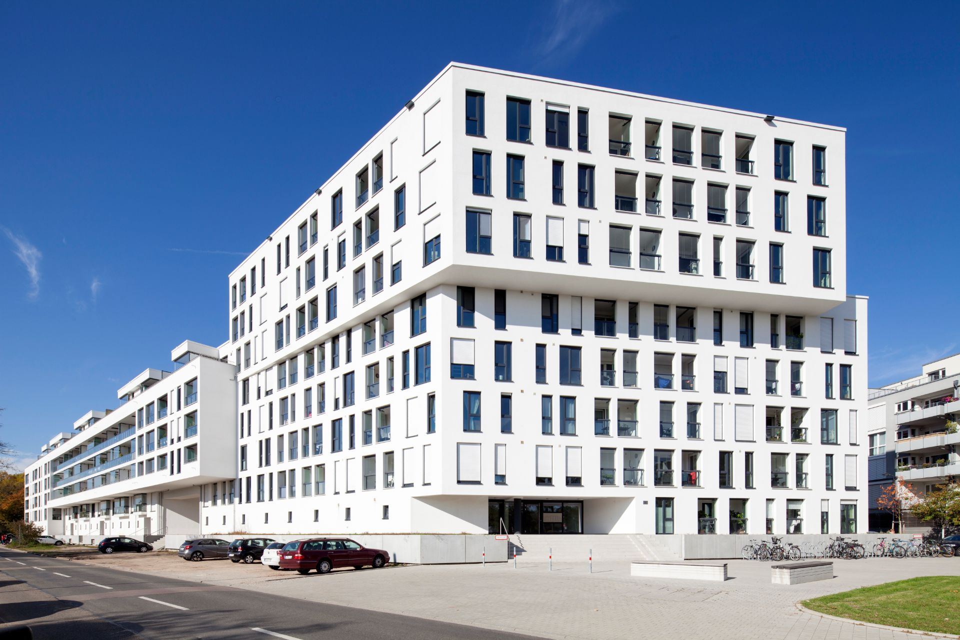 Modernes, mehrstöckiges Wohngebäude mit vielen Fenstern, geparkten Autos und Fahrrädern davor.