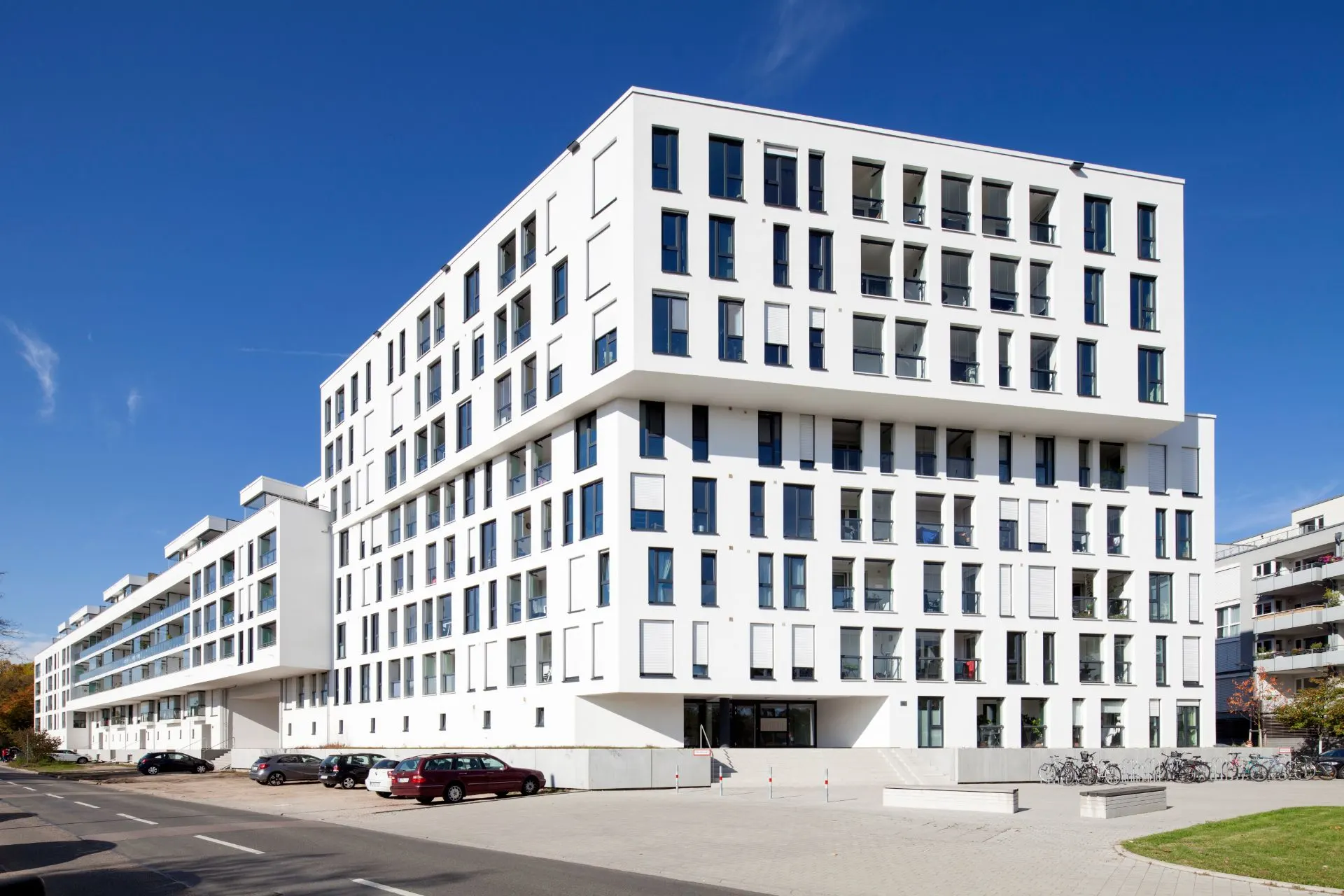 Modernes, mehrstöckiges Wohngebäude mit vielen Fenstern, geparkten Autos und Fahrrädern davor.