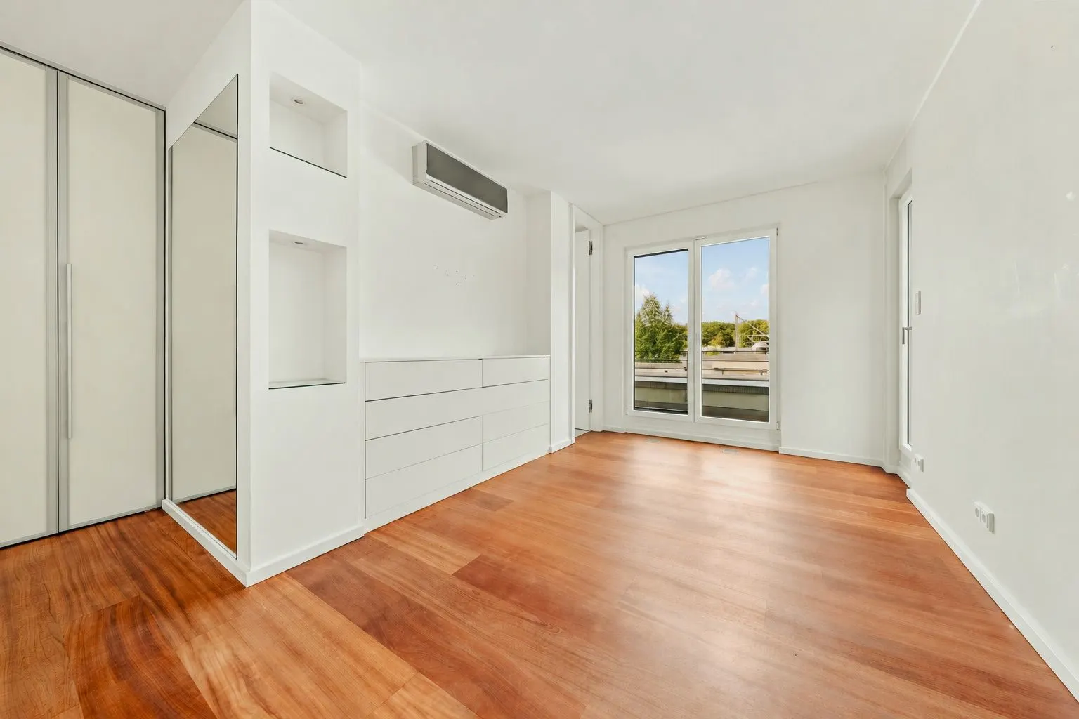 Leeres Zimmer mit Holzfußboden, Einbauschränken und großem Fenster mit Blick nach draußen.