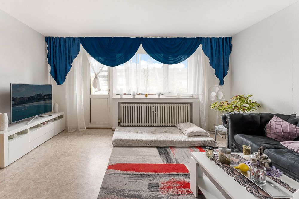 Wohnzimmer mit Sofa, Tisch, Fernseher, Matratze am Fenster und blauen Vorhängen.