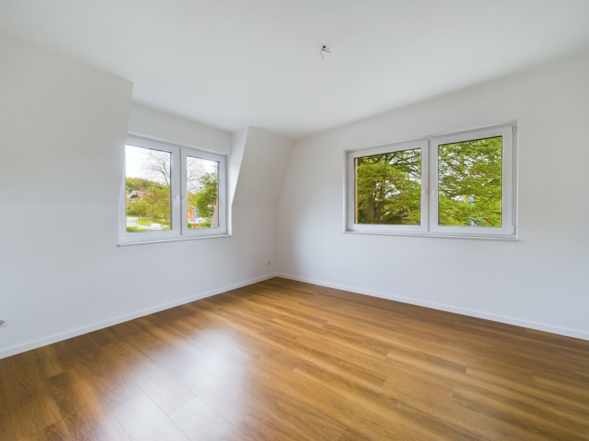 Leerer Raum mit Holzboden und zwei großen Fenstern, weiße Wände.
