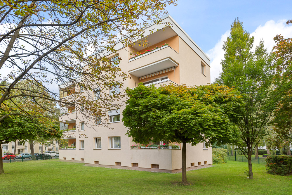 Mehrfamilienhaus mit Balkonen und umgebenden Bäumen auf einer begrünten Wiese.