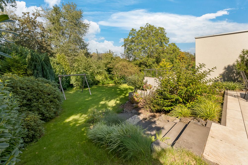 Gepflegter Garten mit Rasenfläche, Schaukelgestell und Terrasse, umgeben von Büschen und Bäumen.