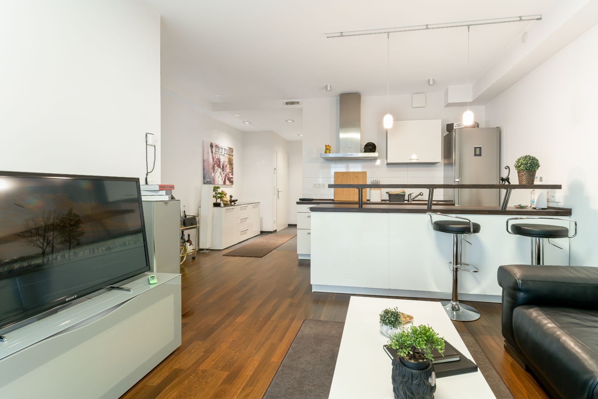 Helles Wohnzimmer mit Fernseher, Sofa, Couchtisch und offener Küche mit Barhockern.