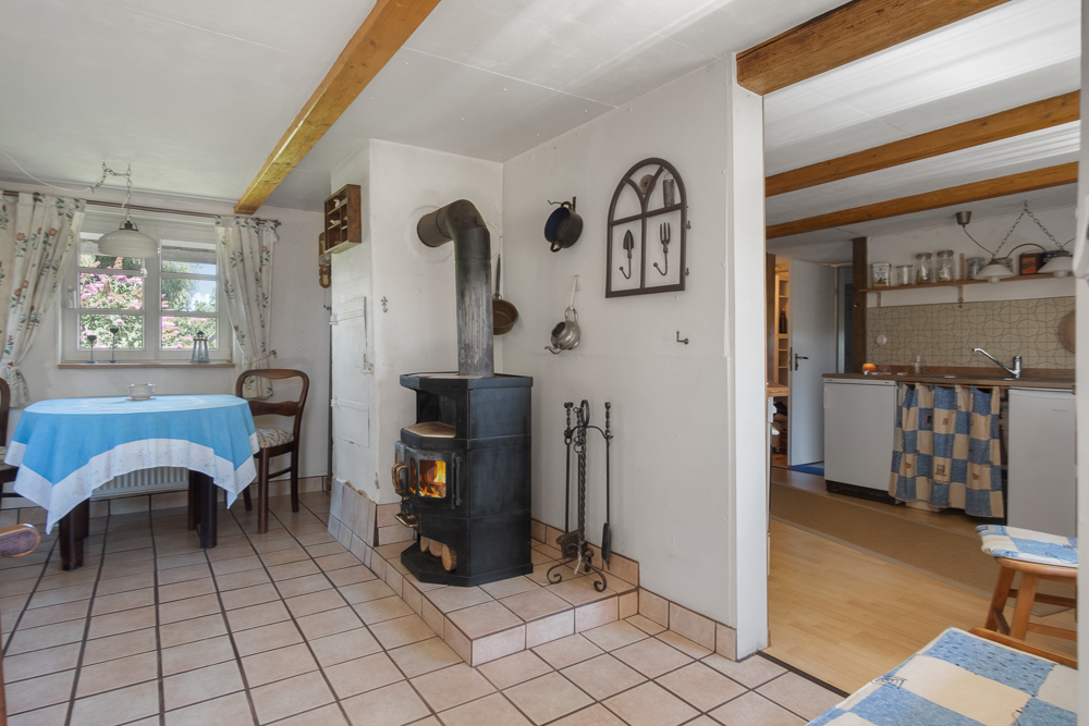 Gemütlicher Wohnbereich mit Esstisch, Holzofen und offener Küche, Fliesenboden und Holzbalken an der Decke.