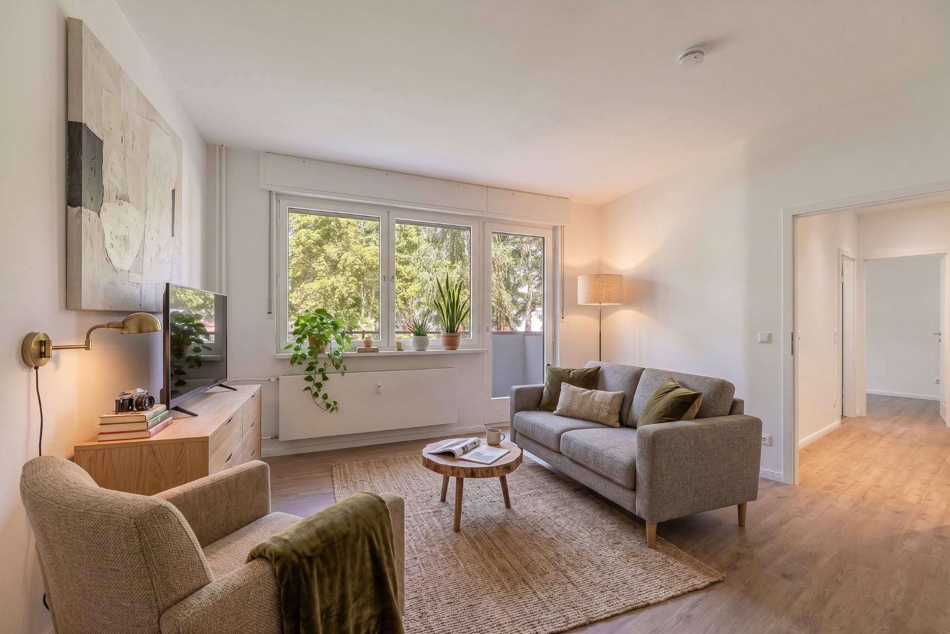 Helles Wohnzimmer mit Sofa, Sessel, Teppich, Couchtisch und Pflanzen vor großen Fenstern.